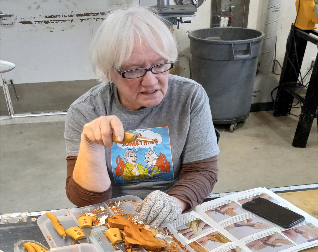 Darlene is holding a woodcarving knife while carving a wooden gold fish.