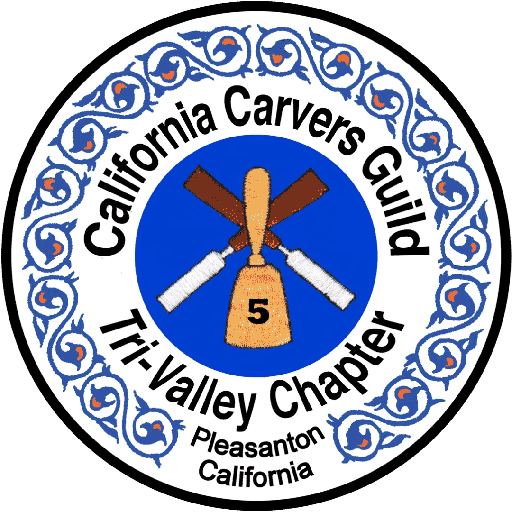 This circular logo of the Tri-Valley Carvers woodcarving club features the name "California Carvers Guild" arched across the top and "Tri-Valley Chapter" curving along the bottom left, set within a white ring bordered by a blue-and-orange decorative vine pattern. At the very bottom center, smaller text reads "Pleasanton, California." The centerpiece is a solid blue circle containing an illustration of three woodcarving tools: two crossed chisels in the background and a single upright mallet with the number 5 on it in the foreground, indicating the chapter is # 5.