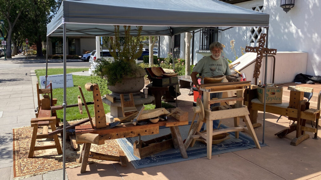 Jim Planting demonstrates woodcarving at the Tri-Valley Carvers Show in Pleasanton, CA, standing behind traditional woodworking benches and an ornate carved cross under a grey canopy.