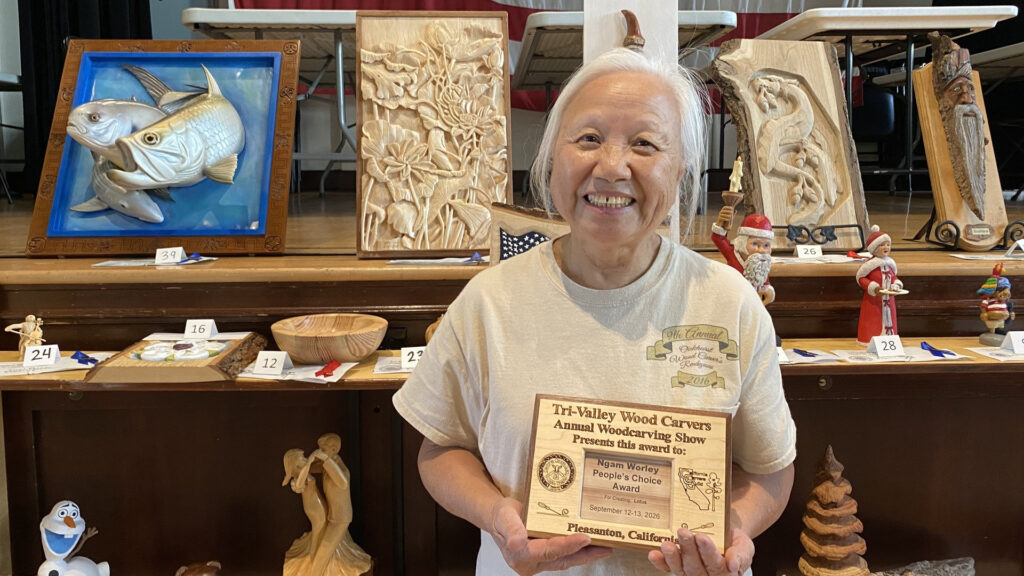 Ngam Worley holds her People's Choice Award for a lotus carving at the Tri-Valley Carvers Show in Pleasanton, CA, standing before a display of her intricate woodcarvings.