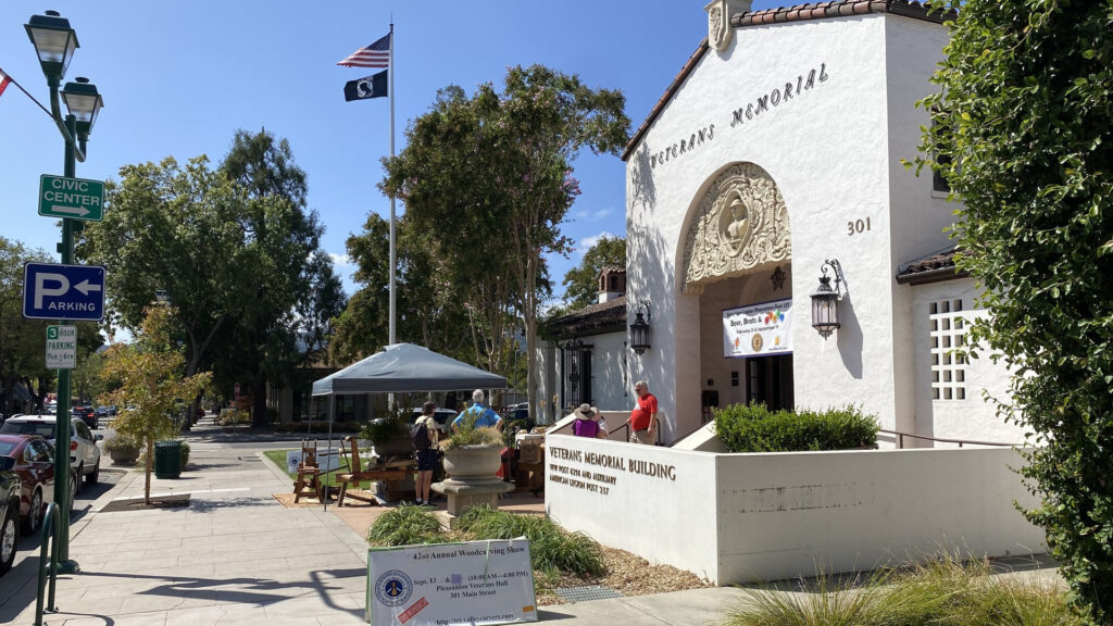 Visitors gather outside the Veterans Memorial Building in Pleasanton, CA, for the Tri-Valley Carvers woodcarving show, featuring a tool display under a grey canopy and an event sign.