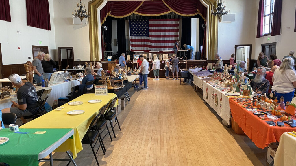 Woodcarvings and craft displays at the Tri-Valley Carvers Show in Pleasanton, CA, featuring a soap-carving station and an American flag backdrop inside the Veterans Memorial Building.