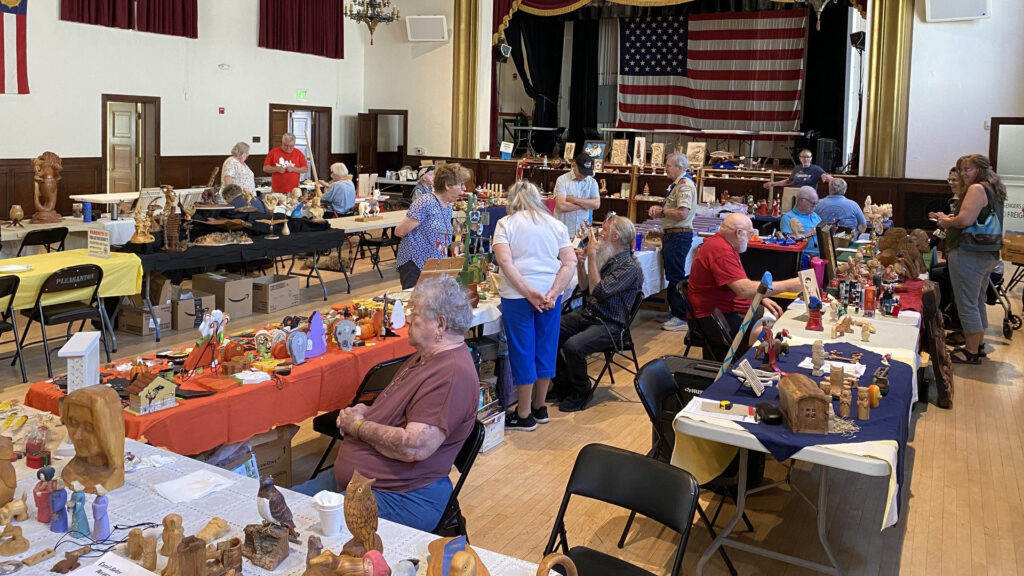 Hand-carved wooden figures, including an owl and a bust, displayed on colorful tables at the 2026 Tri-Valley Carvers Show in Pleasanton, California, with visitors browsing the exhibition.